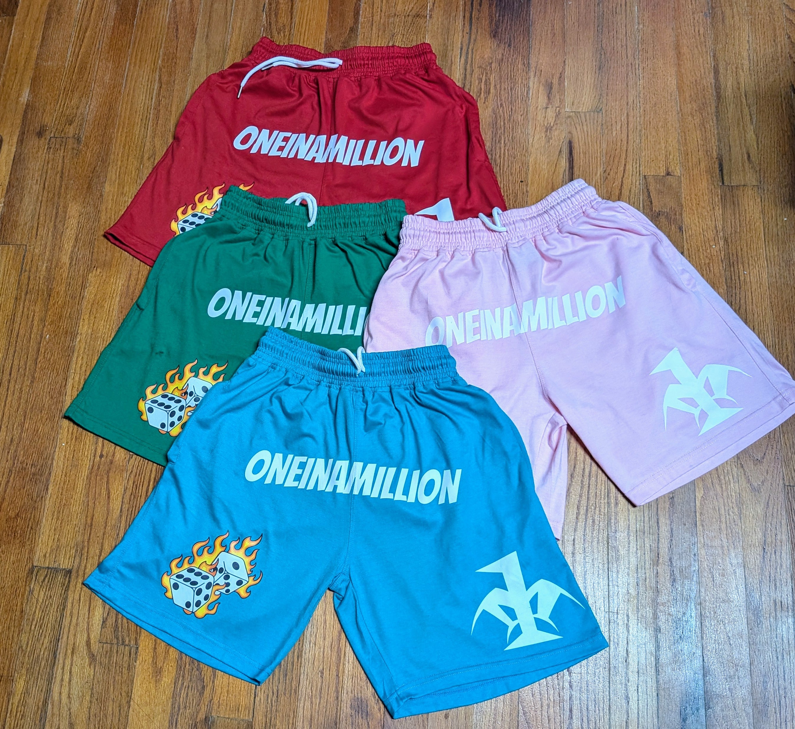 Four pairs of shorts in red, green, blue, and pink with 'ONEINAMILLION' branding on a wooden floor.